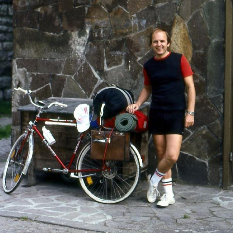 Ein lächelnder Mann steht neben einem stark beladenen Fahrrad vor einer groben Steinwand.