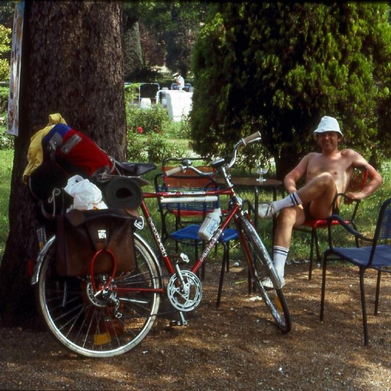 Ein lässig gekleideter Mann posiert neben einem Fahrrad mit Taschen in einer Parklandschaft.