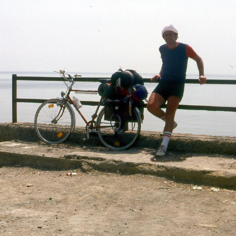 Ein Mann mit Sonnenhut und roten Oberteil posiert mit zwei Fahrrädern bei einer Küstenpromenade.
