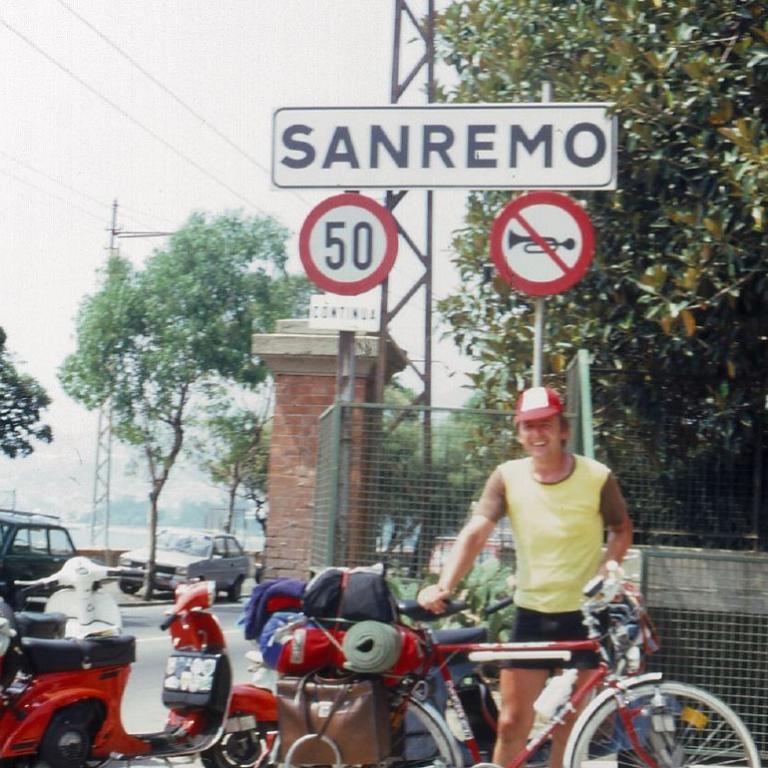 Eine Straße mit einem Eingangsschild nach Sanremo, das von Motorrollern und einem Mann am Bürgersteig belebt wird.