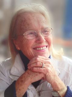 Porträt einer lächelnden älteren Frau mit hellen Haaren, die eine Brille trägt.