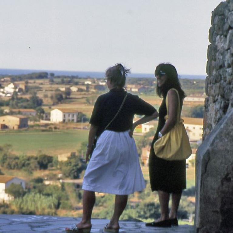 Zwei Frauen stehen vor einer massiven Steinmauer und blicken auf eine weite, mediterrane Landschaft mit Feldern und Häusern.