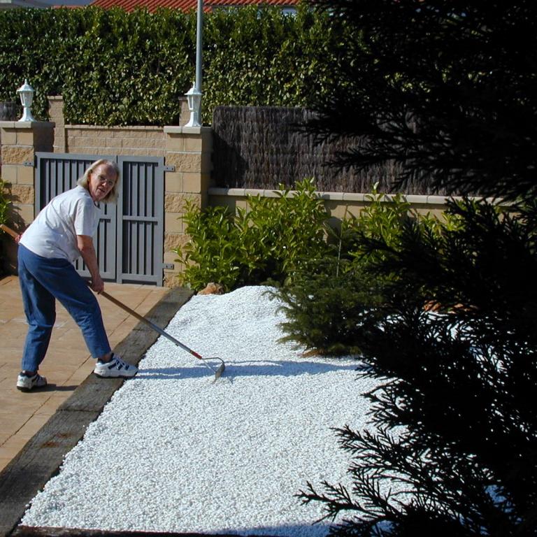 Ein Mann arbeitet im Garten und verteilt weißen Kies vor einer hohen, grünen Hecke.