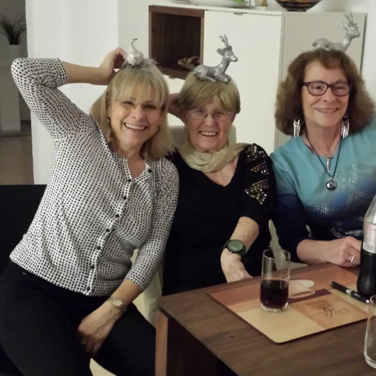 Drei Frauen sitzen an einem Tisch und posieren spielerisch, wobei alle drei auffälligen, dekorativen Kopfaufsätze tragen.