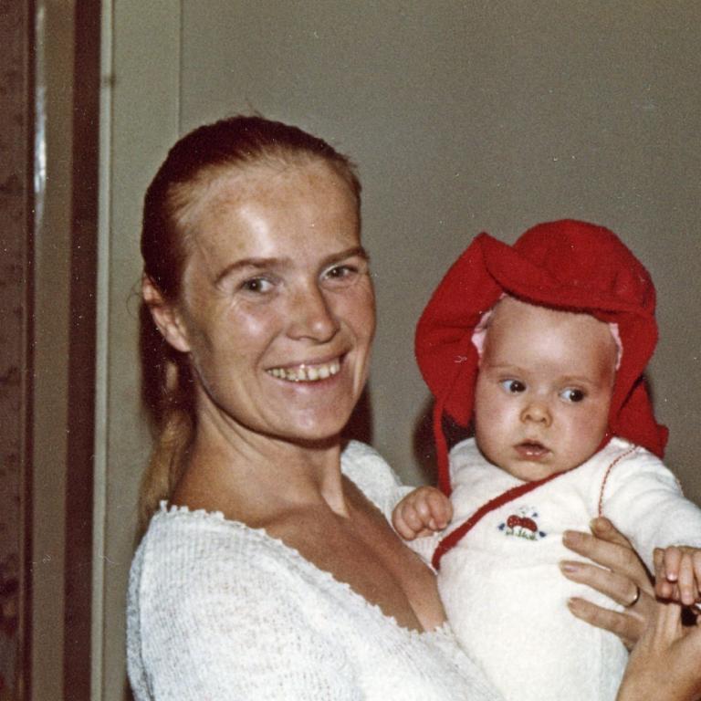 Eine lächelnde Frau hält ein Baby in einem roten Outfit, aufgenommen in einem älteren Stil.