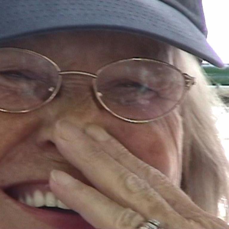 Nahaufnahme einer älteren Frau in Mütze und Brille, die mit ihrer Hand ihr Gesicht bedeckt.