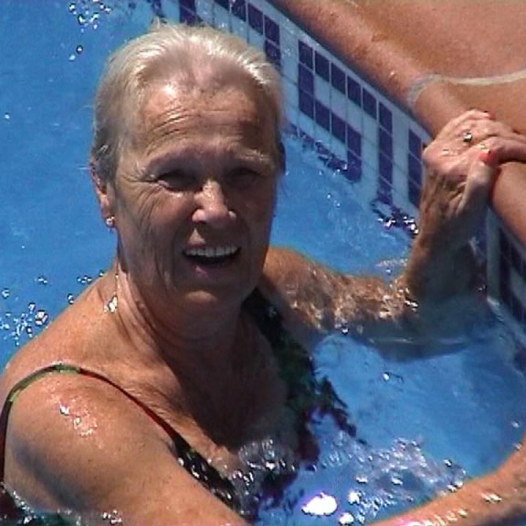 Eine blonde ältere Frau schwimmt lächelnd in einem hellblauen Schwimmbecken.