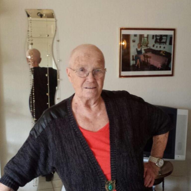 Ein älterer Mann in Brille steht in einem gut beleuchteten Wohnzimmer und trägt einen dunklen Strickcardigan über einem roten Oberteil.