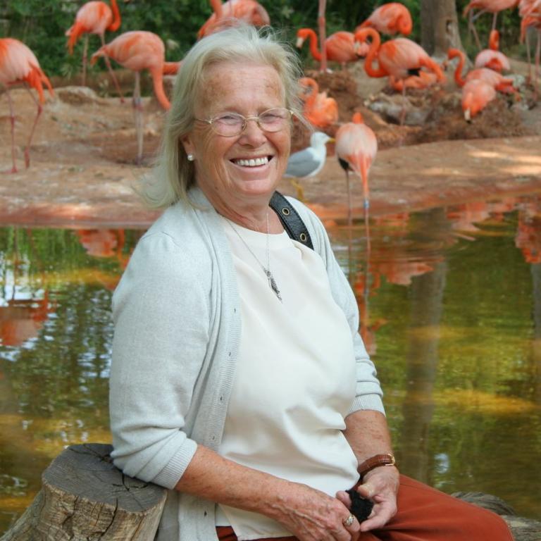Eine lächelnde ältere Frau sitzt am Ufer eines Gewässers mit einer Gruppe Flamingos im Hintergrund.