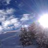 Aufnahme einer verschneiten Berglandschaft mit Nadelbäumen im Vordergrund und einem Seilbahntrack im Hintergrund unter einem strahlend blauen Himmel.