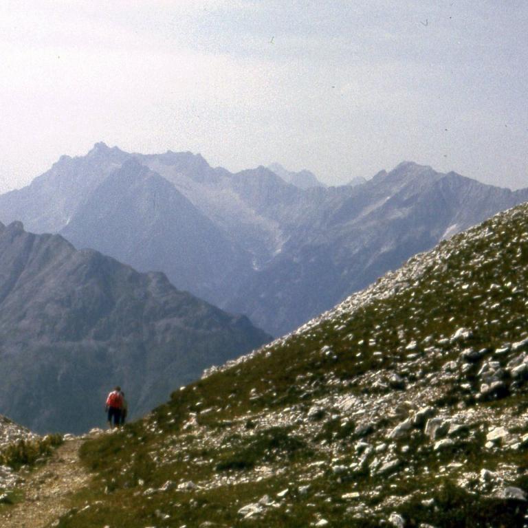 Eine einzelne Person wandert auf einem schmalen Pfad durch eine grasbedeckte Berglandschaft mit Blick auf weitläufige, schichtweise sichtbare Gebirgsz