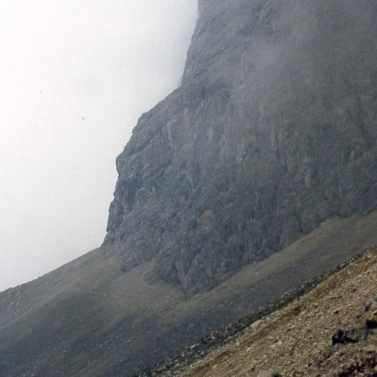Steiler, felsiger Berghang, der teilweise von Nebel verhüllt ist, aufgenommen von einem schrägen, felsigen Hang.