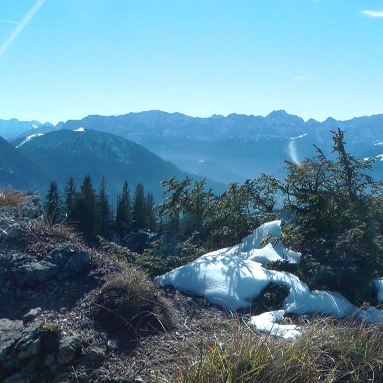 Panoramablick von einem felsigen Bergvorsprung auf eine majestätische Bergkette unter blauem Himmel.