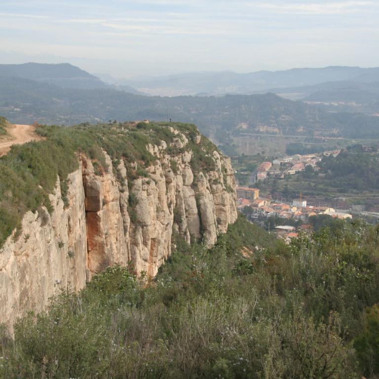 Panoramablick von einer steilen Felsklippe, die auf ein weitläufiges Tal mit grünen Hügeln und Siedlungen blickt.