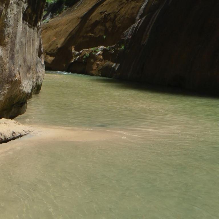 Ein ruhiger Fluss fließt zwischen steilen, hohen Felswänden, und ein kleiner Sandstrand ist am Ufer sichtbar.