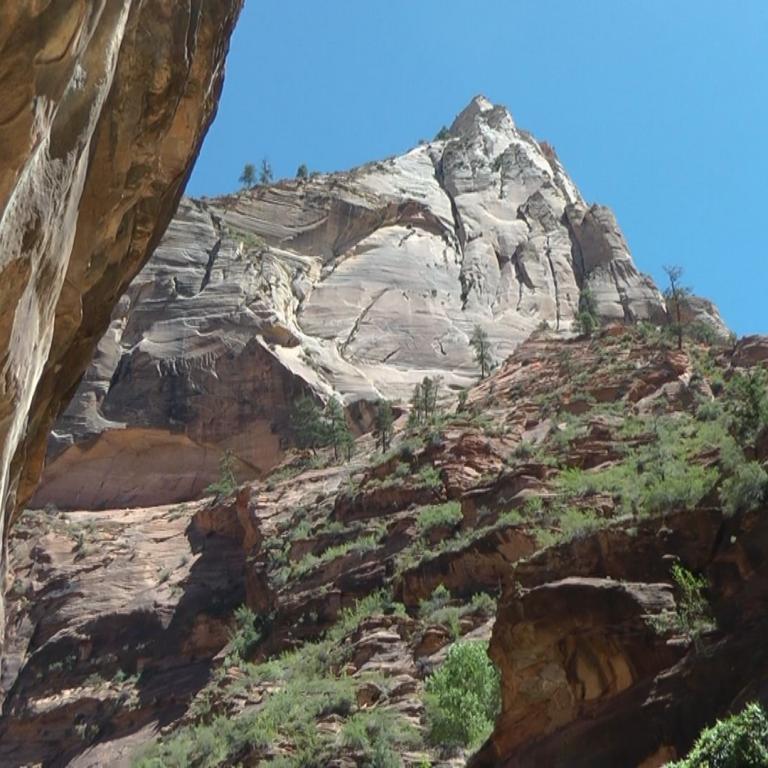 Ein dramatischer Blick auf eine steile Felsformation mit massiven Canyonwänden im Vordergrund unter einem klaren blauen Himmel.